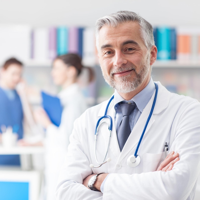 May include: A smiling doctor with gray hair and a beard, wearing a white coat, blue tie, and stethoscope. The doctor has their arms crossed. Other people are blurred in the background.