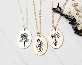 May include: Three silver, gold, and rose gold necklaces with oval pendants featuring delicate line drawings of flowers. The flowers are a lily of the valley, a daffodil, and a poinsettia.
