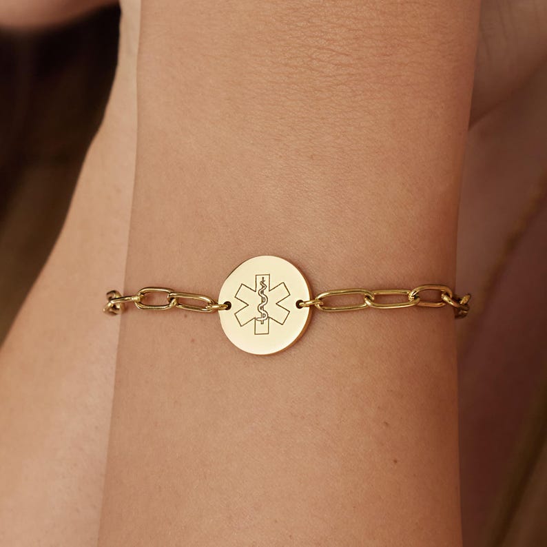 May include: Gold-toned medical alert bracelet. The bracelet features a chain link design with a circular pendant. The pendant displays the Star of Life symbol, a six-pointed star with a serpent-entwined staff in the center, a universal symbol for emergency medical services.