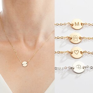 May include: A gold chain necklace with a small round pendant featuring a heart design. The necklace is shown on a model. Three other necklaces are shown below, each with a different pendant: a letter M, a paw print, and a letter B.