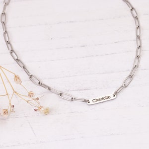 May include: A silver-colored chain necklace with a rectangular pendant engraved with the name "Charlotte." The necklace features a link chain design. The background is a white wooden surface with a few small, dried flowers.