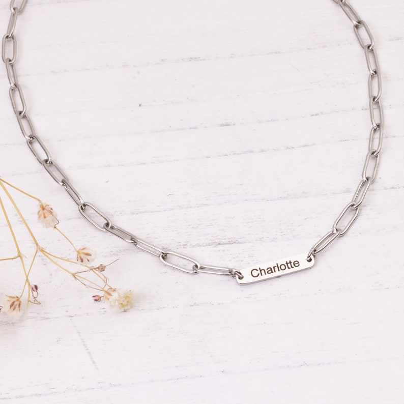 May include: A silver-colored chain necklace with a rectangular pendant engraved with the name "Charlotte." The necklace features a link chain design. The background is a white wooden surface with a few small, dried flowers.