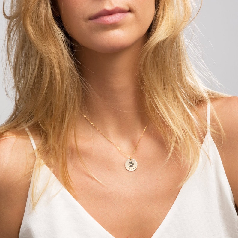 May include: A gold necklace with a small round pendant featuring a paw print. The pendant is on a delicate chain.