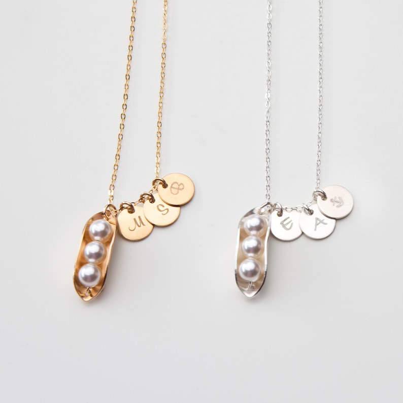 Puede incluir: Dos collares con colgantes de vaina de guisante y colgantes de letras personalizados. El collar de la izquierda tiene una cadena dorada y colgantes dorados con las letras "M", "S" y "B". El collar de la derecha tiene una cadena plateada y colgantes plateados con las letras "W", "A" y un s&iacute;mbolo de ancla n&aacute;utica.