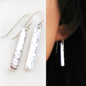 May include: Silver hammered bar earrings with a textured finish. The earrings are hanging from a silver hook.