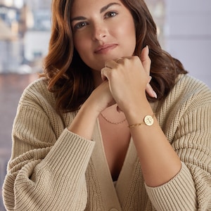 May include: A gold-colored bracelet with a circular charm featuring a design is worn on a wrist. The person is wearing a beige cardigan and a delicate necklace. The background shows a blurred cityscape.