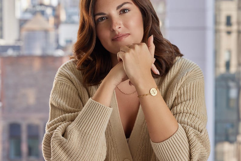 May include: A gold-colored bracelet with a circular charm featuring a design is worn on a wrist. The person is wearing a beige cardigan and a delicate necklace. The background shows a blurred cityscape.