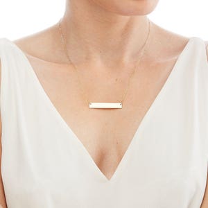 May include: A gold bar necklace with a simple, flat bar pendant. The necklace is worn by a person with a white top.