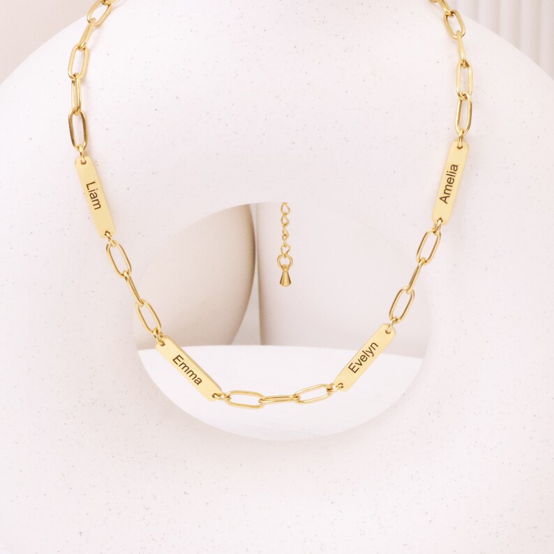 May include: A gold-colored necklace with a chain link design. The necklace features four rectangular pendants, each engraved with a name: Liam, Emma, Evelyn, and Amelia. The necklace is displayed against a white background.
