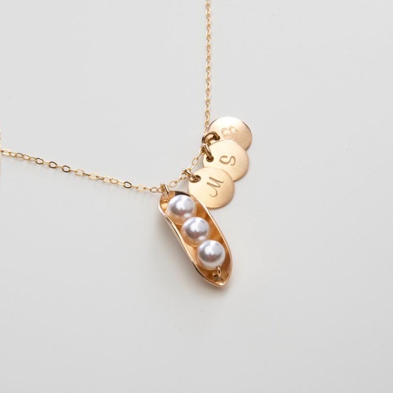 Puede incluir: Collar de oro con un dije en forma de vaina de guisante que contiene tres perlas y tres discos de oro grabados con las letras 'M', 'S' y 'B'.