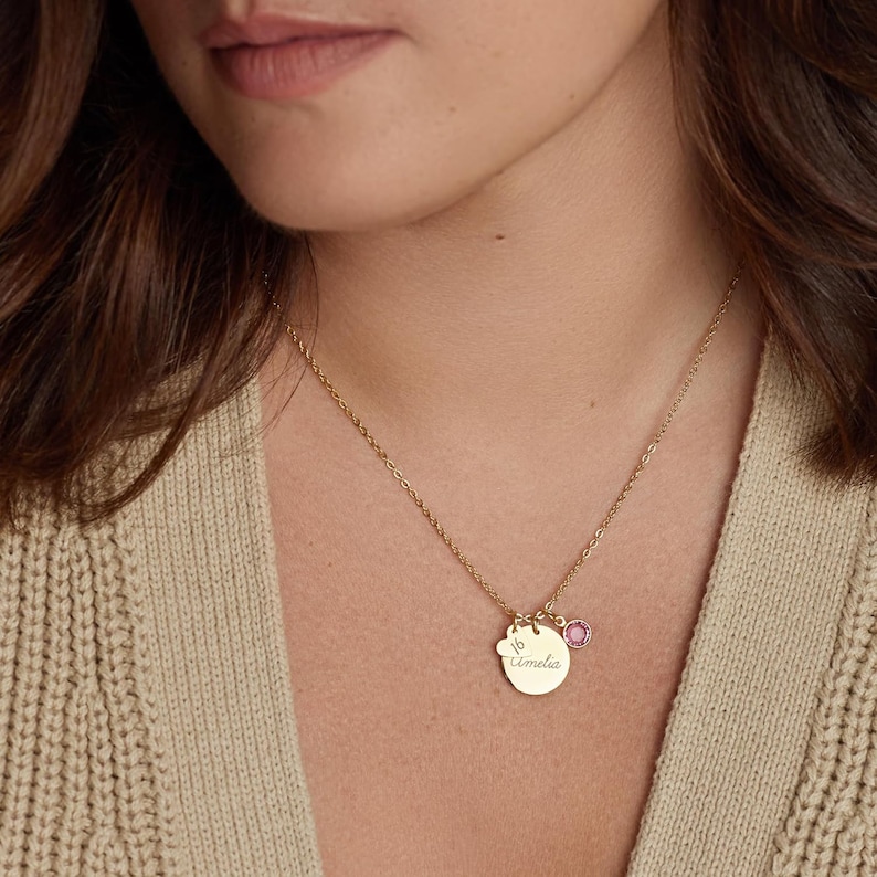 May include: A gold-toned necklace featuring a circular pendant engraved with the name Amelia and the number 16, accompanied by a small, round, pink-coloured gemstone charm. The pendant and charm hang from a delicate gold-toned chain.