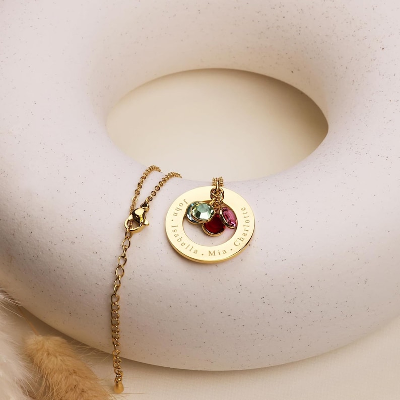 May include: Gold necklace with a round pendant engraved with the names "Mia", "Isabella", "Maya", and "Charlotte". The pendant has four colorful gemstones attached.