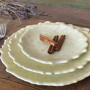 May include: A stack of three pale yellow, scalloped-edge plates with a floral pattern. Cinnamon sticks rest on the top plate. A silver fork and dried lavender sprigs are also present, set on a wooden surface.