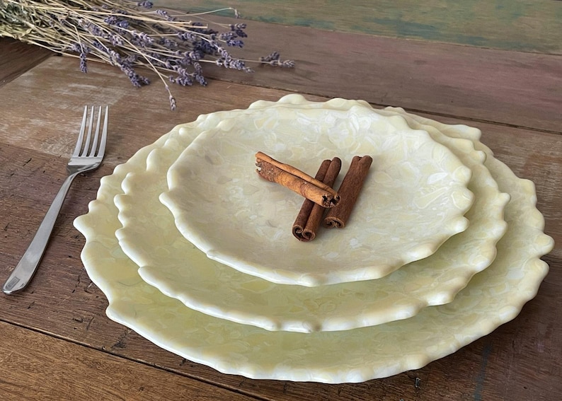 May include: A stack of three pale yellow, scalloped-edge plates with a floral pattern. Cinnamon sticks rest on the top plate. A silver fork and dried lavender sprigs are also present, set on a wooden surface.