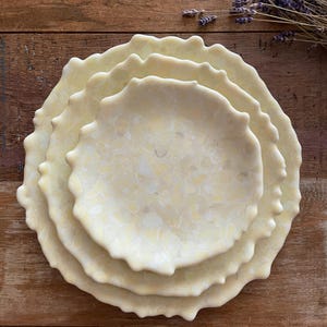 May include: A stack of four off-white ceramic plates with a textured, wavy edge. The plates have a marbled pattern in shades of cream and pale yellow. The plates are arranged on a wooden surface, with a sprig of dried lavender in the background.