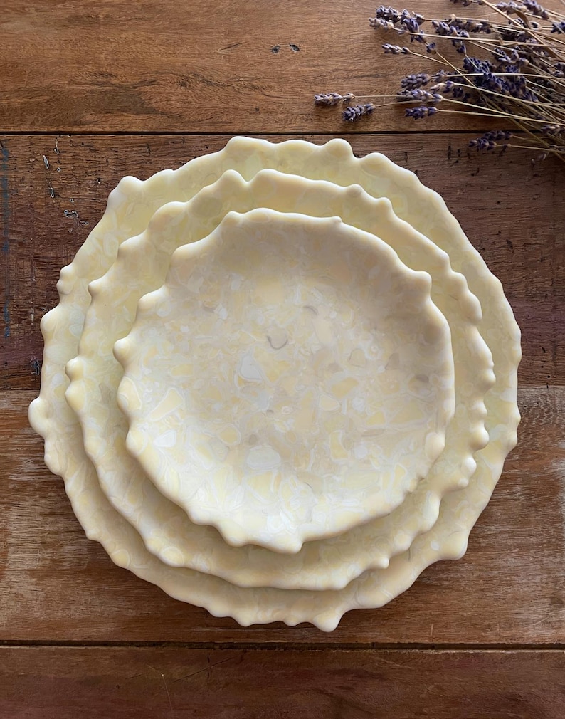 May include: A stack of four off-white ceramic plates with a textured, wavy edge. The plates have a marbled pattern in shades of cream and pale yellow. The plates are arranged on a wooden surface, with a sprig of dried lavender in the background.