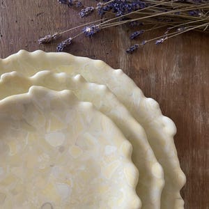 May include: A stack of three off-white ceramic plates with a marbled pattern and scalloped edges. Dried lavender sprigs rest on a wooden surface in the background. The plates have a diameter of approximately 10 inches.