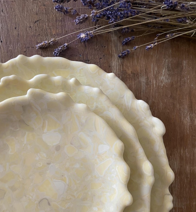 May include: A stack of three off-white ceramic plates with a marbled pattern and scalloped edges. Dried lavender sprigs rest on a wooden surface in the background. The plates have a diameter of approximately 10 inches.