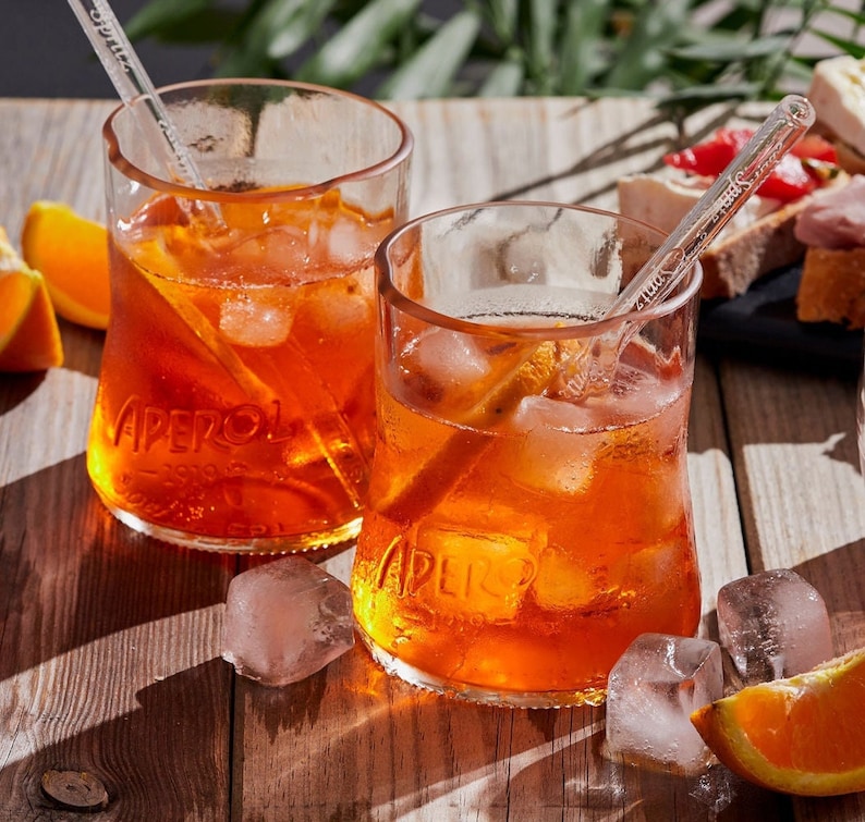 May include: Two glasses of Aperol Spritz with ice cubes and straws on a wooden table. The glasses are filled with a bright orange liquid and have the Aperol logo printed on them. There are orange slices and ice cubes on the table.