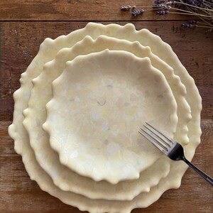 May include: A stack of four cream-colored ceramic plates with a wavy, textured edge. A silver fork rests on the plates. The plates have a marbled pattern and are arranged on a wooden surface. Dried lavender sprigs are in the background.