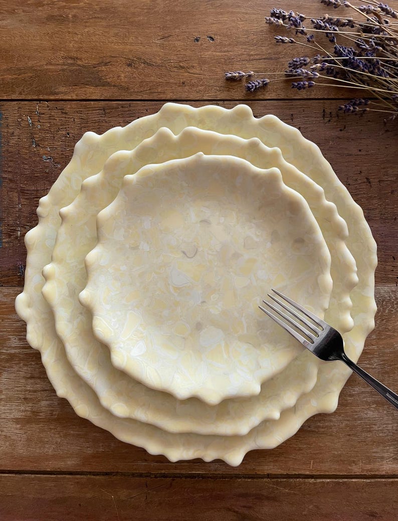 May include: A stack of four cream-colored ceramic plates with a wavy, textured edge. A silver fork rests on the plates. The plates have a marbled pattern and are arranged on a wooden surface. Dried lavender sprigs are in the background.