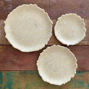 May include: Three off-white, round ceramic plates with a textured surface and scalloped edges. The plates are arranged on a wooden surface with a mix of brown, red, and green tones.