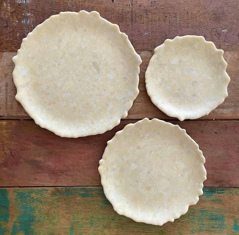 May include: Three off-white, round ceramic plates with a textured surface and scalloped edges. The plates are arranged on a wooden surface with a mix of brown, red, and green tones.