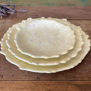 May include: A stack of three decorative, scalloped-edge plates in varying sizes. The plates have a creamy, speckled appearance with a light yellow hue. The plates are arranged on a wooden surface.