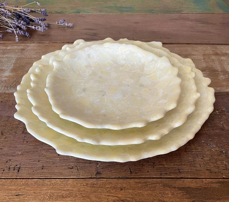 May include: A stack of three decorative, scalloped-edge plates in varying sizes. The plates have a creamy, speckled appearance with a light yellow hue. The plates are arranged on a wooden surface.