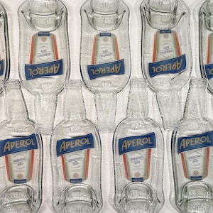 Puede incluir: Botellas y bandejas de servicio de vidrio transparente de Aperol. Las botellas y bandejas tienen el logotipo de Aperol en azul y blanco. El logotipo de Aperol está en una pancarta azul con la palabra "APEROL" en blanco. Las bandejas tienen forma de botella de Aperol.