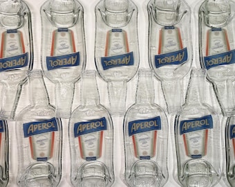 Może przedstawiać: Przezroczyste szklane butelki Aperol i tace do serwowania w kształcie butelek. Butelki i tace mają logo Aperol w kolorze niebieskim i białym. Logo Aperol znajduje się na niebieskim banerze ze słowem "APEROL" w kolorze białym. Tace mają kształt butelki Aperol.