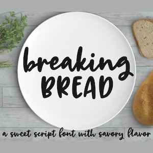 Könnte beinhalten: Ein weißer Teller mit dem Text "breaking BREAD" in schwarzer, handschriftlicher Schrift. Der Text befindet sich auf einem weißen Teller, der auf einer Holzoberfläche liegt. Auf der Oberfläche befinden sich zwei Brote.