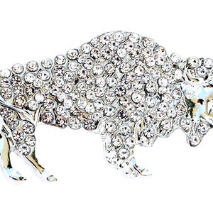 May include: A silver-toned metal brooch shaped like a buffalo, entirely covered in clear, sparkling round rhinestones. The animal figure is depicted in profile, showcasing the distinct hump, head, and legs, with a polished metallic finish visible.