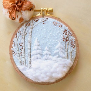 Peut inclure: Une broderie en forme de cercle représentant une scène d'hiver enneigée avec des arbres et une épaisse couche de neige blanche. Le cercle est en bois et possède un crochet en métal doré.