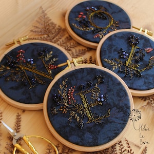 Peut inclure: Quatre cerceaux brodés bleu marine avec des motifs floraux dorés et perlés. Chaque cerceau présente une lettre différente, avec des broderies dorées et des accents perlés dans des tons de rouge, bleu et or. Les cerceaux sont exposés sur une surface brun clair.