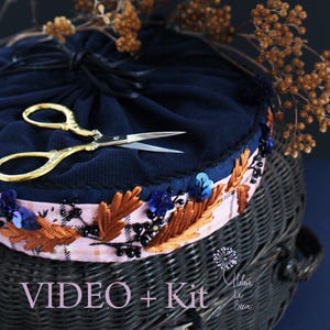 Peut inclure: Une pochette en tissu bleu marine avec fermeture à cordon, ornée d'une bande rose pâle brodée de feuilles marron, de perles noires et de sequins bleus. Une paire de ciseaux de broderie dorés est posée dessus. Le texte indique VIDEO + Kit.