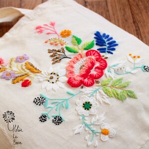 Könnte beinhalten: Eine weiße Canvas-Tragetasche mit bunten gestickten Blumen. Die Blumen sind in den Farben Rosa, Rot, Gelb, Grün, Blau und Weiß gehalten. Die Tasche hat ein schwarzes gesticktes Design mit dem Schriftzug "Hélene Le Bern" am Boden.