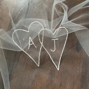 May include: Two white embroidered hearts on a white tulle veil. The hearts are intertwined and have the initials "A" and "J" stitched inside.