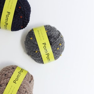 May include: Three balls of yarn with yellow and black polka dots. The yarn is in three colors: navy blue, gray, and brown. Each ball has a yellow label that says "Pom Pom" in black letters.
