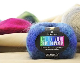 Puede incluir: Primer plano de una madeja de lana azul y morada con una etiqueta que dice "EXCELLENT MOHAIR (COUNT 10) GRADATION". Se ven otras madejas de lana en tonos rosa, verde y gris en el fondo. La lana es suave y esponjosa.
