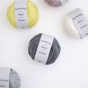 Puede incluir: Cinco ovillos de lana en amarillo, blanco, gris oscuro, gris claro y malva, cada uno envuelto en una banda de papel gris claro. Las bandas tienen impreso el texto Rambouillet Wool Cotton. El hilo está dispuesto de forma dispersa sobre una superficie lisa.