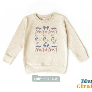 May include: A beige sweatshirt with a pattern of ducks, hearts, and flowers. The ducks are wearing blue bows. The text "Ships Next Day" is visible at the bottom of the image.