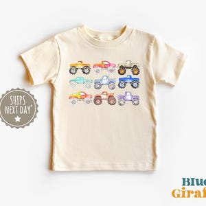 May include: A cream colored t-shirt with a colorful print of monster trucks. The trucks are in various shades of blue, green, red, orange, and yellow.