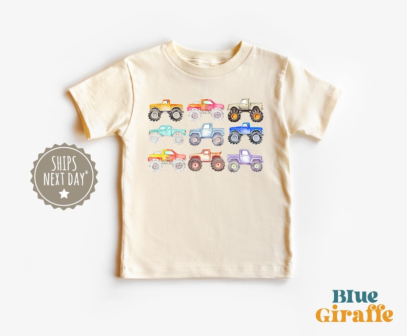 May include: A cream colored t-shirt with a colorful print of monster trucks. The trucks are in various shades of blue, green, red, orange, and yellow.