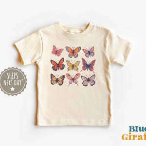 May include: A cream-colored short-sleeve t-shirt featuring a graphic print of nine butterflies in various colors including orange, yellow, pink, and blue. The butterflies are arranged in a three-by-three grid pattern on the center of the chest.