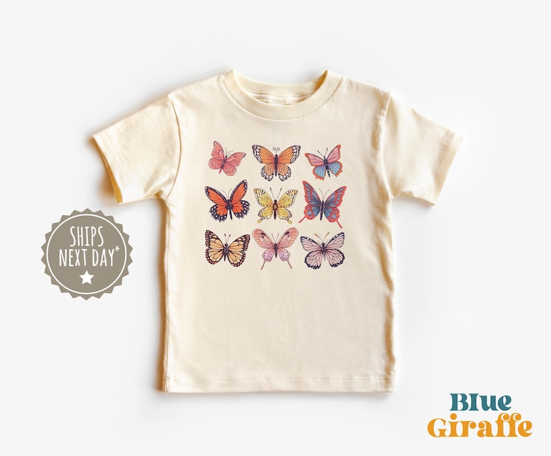 May include: A cream-colored short-sleeve t-shirt featuring a graphic print of nine butterflies in various colors including orange, yellow, pink, and blue. The butterflies are arranged in a three-by-three grid pattern on the center of the chest.