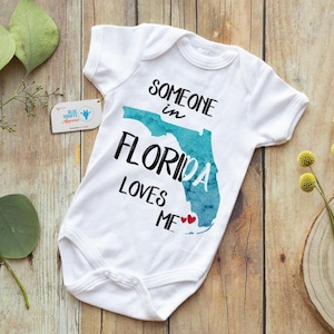 May include: White baby onesie with the text "SOMEONE in FLORIDA LOVES ME" and a teal Florida state outline. Two red hearts are below the text. The onesie is on a wooden surface.