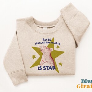 May include: A beige sweatshirt with a graphic of a rat inside a green star, with the text "RATS SPELLED BACKWARDS IS STAR". The sweatshirt has long sleeves and a crew neck.