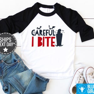 May include: A white and black baseball tee shirt with a red and blue graphic that says "Careful I Bite" with a vampire silhouette and bats.