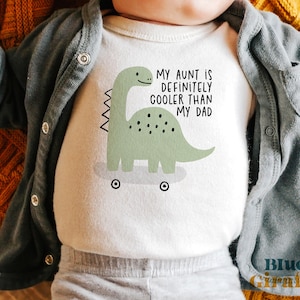 Könnte beinhalten: Weißer Baby-Body mit einem grünen Dinosaurier-Motiv und dem Text "MY AUNT IS DEFINITELY COOLER THAN MY DAD". Der Dinosaurier hat Räder. Das Baby trägt eine graue Strickjacke und eine Hose.